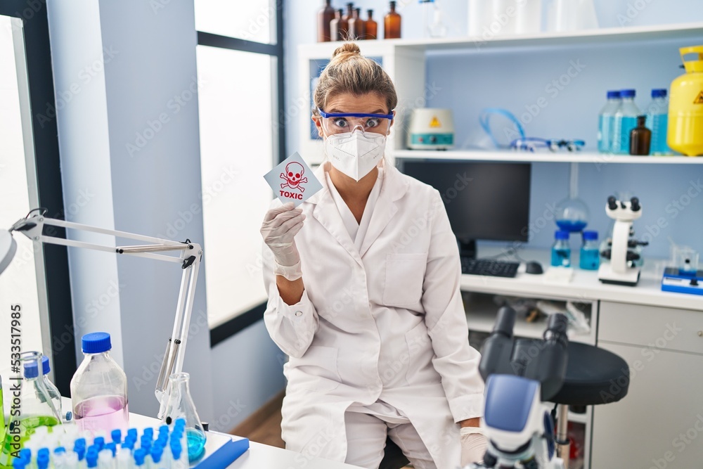 Sticker young woman working at scientist laboratory holding toxic banner scared and amazed with open mouth f