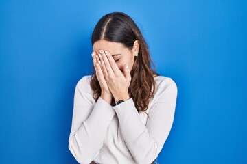 Young hispanic woman standing over blue background with sad expression covering face with hands...