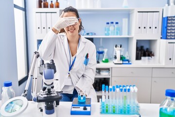 Young hispanic woman working at scientist laboratory smiling and laughing with hand on face covering eyes for surprise. blind concept.