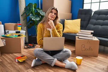 Young woman sitting on the floor at new home using laptop with hand on chin thinking about question, pensive expression. smiling with thoughtful face. doubt concept.