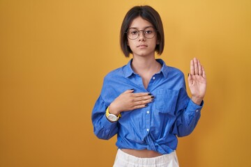 Young girl standing over yellow background swearing with hand on chest and open palm, making a loyalty promise oath