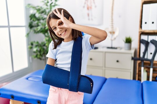 Young hispanic girl wearing arm on sling at rehabilitation clinic smiling happy doing ok sign with hand on eye looking through fingers