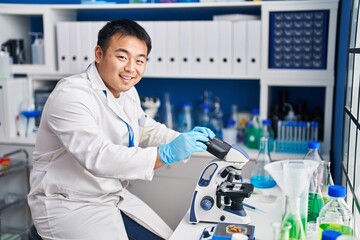 Young chinese man wearing scientist uniform using microscope at laboratory