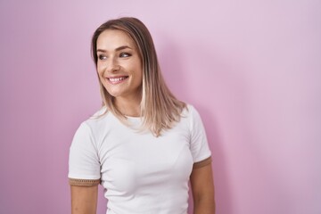 Blonde caucasian woman standing over pink background looking away to side with smile on face, natural expression. laughing confident.
