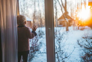 little boy looks out the window at home in winter waving his hand and feeling sad on a cold day