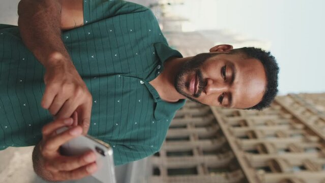 VERTICAL VIDEO: Young man standing on the street next to the road and using the phone