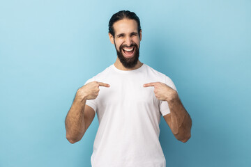Portrait of delighted man with beard wearing white T-shirt pointing fingers on himself and looking...