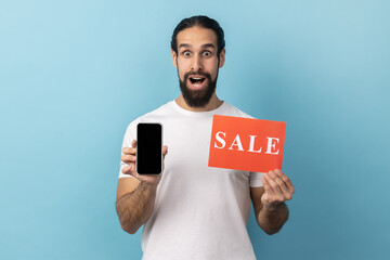 Portrait of man with beard wearing white T-shirt widely open eyes and mouth holding sale card and smartphone, shocked with amazing online sell-out. Indoor studio shot isolated on blue background.