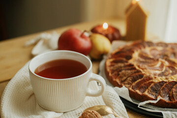 Warm tea in stylish cup on rustic wooden table with freshly baked apple pie with cinnamon, candle, autumn leaves,  nuts. Autumn hygge still life. Atmospheric cozy home
