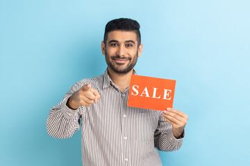 Portrait of smiling satisfied businessman pointing at camera and holding card with sale inscription, low prices on Black Friday, wearing striped shirt. Indoor studio shot isolated on blue background.