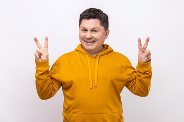 Fototapeta premium Happy positive middle aged man showing v sign to camera, winking and smiling, male wishing good luck, peace, wearing urban style hoodie. Indoor studio shot isolated on white background.