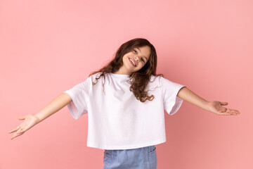 Portrait of positive optimistic little girl wearing white T-shirt inviting to embrace, keeping hands as giving, sharing for free. Indoor studio shot isolated on pink background.