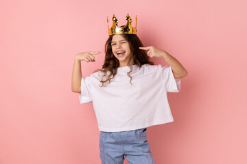 Portrait of happy confident smiling little girl wearing white T-shirt standing in gold crown, pointing at herself with pride, looking at camera. Indoor studio shot isolated on pink background. - obrazy, fototapety, plakaty