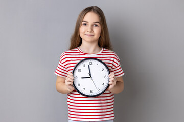 Portrait of smiling little girl wearing striped T-shirt holding big wall clock, looking at camera...