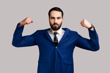Strong confident bearded man raising hands showing power, feeling independent strong with proud look, wearing official style suit. Indoor studio shot isolated on gray background.