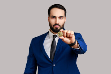 Handsome bearded man holding out gold bitcoin, looking at camera with toothy smile, blockchain concept, wearing official style suit. Indoor studio shot isolated on gray background.