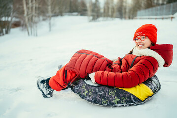 a teenage girl in a red jacket in a snowy forest rides in a tubing on a cold day