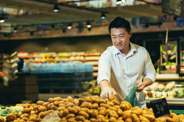 A young handsome Asian man picks potatoes in a green plastic bag in a supermarket in the vegetable department.