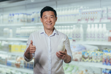 Advertising. Portrait of a young handsome Asian man standing in a supermarket at the dairy department and holding a plastic bottle of milk in his hands. He points to OK, smiles, looks at the camera.
