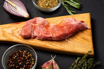 Raw meat steaks on a cutting board. Dark background. Preparation for cooking pork meat. Various spices, seasonings lie nearby.