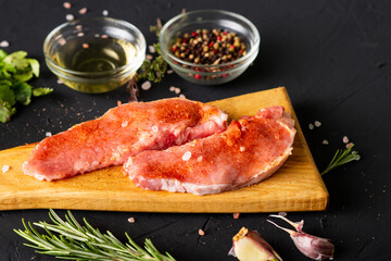 Raw meat on a cutting board. Dark background. Preparation for cooking pork meat. Various spices, seasonings lie nearby.
