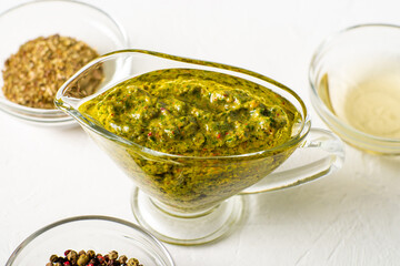 Chimichurri sauce in a gravy bowl on a white background. Various spices lie nearby. Argentinean vegetarian sauce made from olive oil, oregano, parsley.