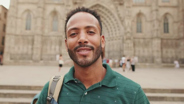 Close-up of young man tourist with backpack on his shoulder, looking at the camera and smiling while standing on the square of the old city