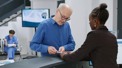 Fototapeta premium Senior man paying with credit card at hospital reception desk, making electronic payment transaction for medical checkup appointment and treatment. Receptionist helping man to pay.