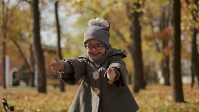 Happy cute brown-haired girl with down syndrome in fashionable coat and stylish eyeglasses tossing bright foliage up to the sky and laughing, kid enjoying time in warm autumn park, happy childhood