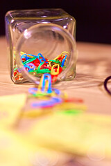 Square jar on its' side pouring paperclips and push pins onto a wooden table with sticky notes and other stationary. Looking inside the jar with a shallow depth of field, blurred bokeh. Productivity.