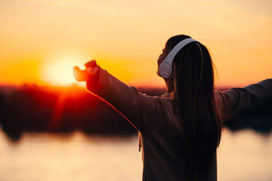 Young woman with outstretched arms standing on a hill above the river watching the sunset while listening to music via headphones