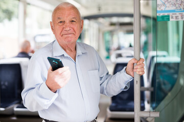 Old Caucasian man with smartphone standing in streetcar and waiting for next stop.