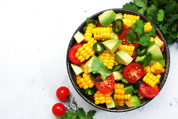 Mexican spicy salad with corn, avocado, jalapeno peppers, cherry tomatoes and cilantro. White kitchen table background, top view