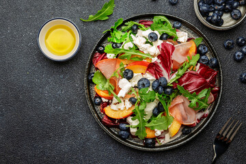 Fresh salad with arugula, radicchio, sweet peaches, smoked pork ham, feta cheese and blueberries. Black stone table background, top view