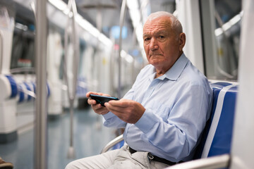 Senior Caucasian man with smartphone sitting on seat in subway car.