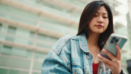 Close up long haired Asian girl texting on smartphone waiting friend outdoors. Beautiful woman dressed casual using mobile phone on street