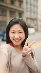 Pretty Asian girl taking self portrait showing peace sign outdoors. Long haired smiling woman posing on street