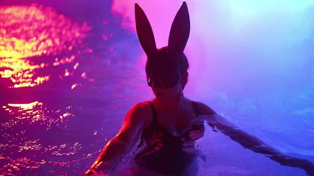 Sexy woman in bunny latex mask swimming in pool at night. Attractive chick in black swimsuit enjoying party time. Rich lifestyle, luxury life, entertainment, masquerade. Dangerous girl