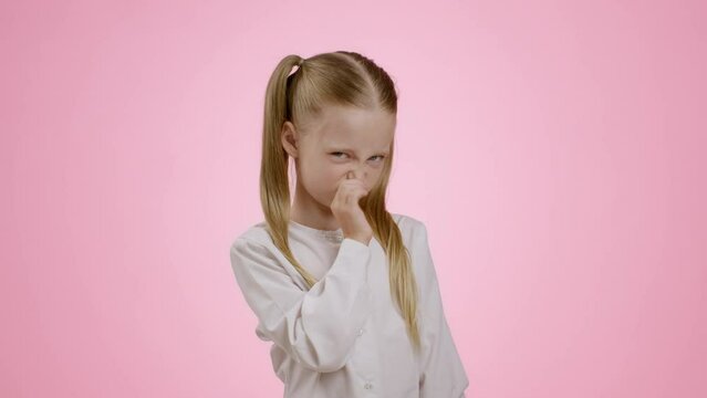 Awful odor. Cute little girl smelling something terrible, closing her nose and grimacing, frowning face, pink background