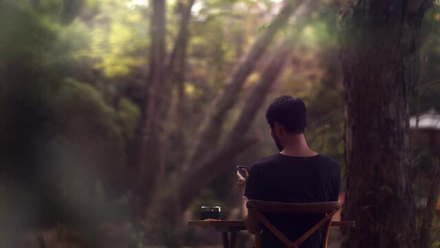 asian man sitting outside in the garden at a table and working on his smartphone - cinematic shot