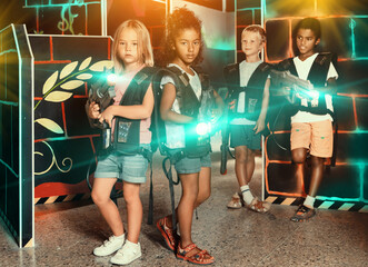 Two preteen girls having fun with friends on the dark gaming arena, posing back to back with laser guns