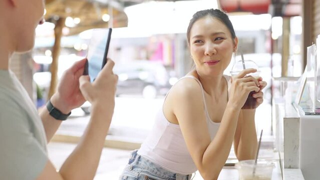 Asian couple enjoy and fun outdoor lifestyle shopping at street market on summer holiday vacation. Handsome man using mobile phone photography his girlfriend while shopping together at weekend market