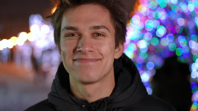 Headshot portrait of satisfied carefree Caucasian boy looking at camera smiling admiring urban city illumination on Christmas eve. Close-up of happy smiling guy posing outdoors at New Year night