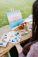 Unrecognizable young female artist painting her own beach scenery with oil on a canvas outdoor....
