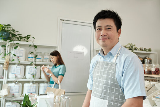 Portrait of an Asian male shopkeeper smiling and looking at camera, arranges natural products at refill store, zero-waste grocery, and plastic-free, eco environment-friendly, sustainable lifestyles.