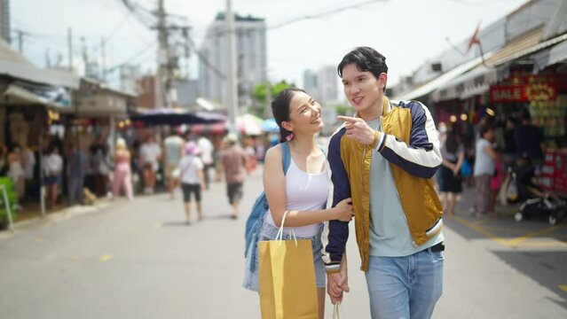 4K Attractive Asian couple enjoy and fun outdoor lifestyle shopping at street market while travel on summer holiday vacation. Happy man and woman couple holding shopping bag walking at weekend market