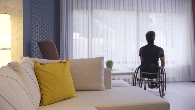 Young man sitting in wheelchair walking around the house and looking out the window.
Disabled young man is thoughtful and looks out the window of the house.
