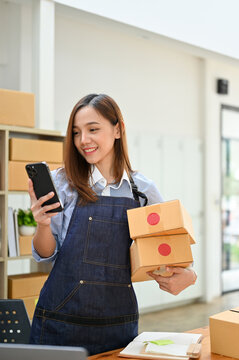 Portrait, Asian Female Small Online Business Owner Holding Shipping Boxes And Using Her Phone