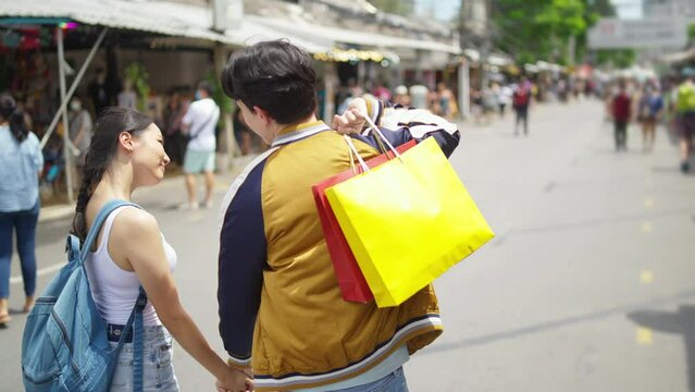 4K Attractive Asian couple enjoy and fun outdoor lifestyle shopping at street market while travel on summer holiday vacation. Happy man and woman couple holding shopping bag walking at weekend market
