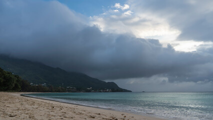 Fototapeta na wymiar A foggy day on a tropical beach. The turquoise ocean is calm. Foam of waves on the sand. A rain cloud hung over the hill. Seychelles. Mahe. Beau Vallon